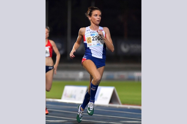 Lucy Evans running on an athletics tracks. Lucy is a white woman with dark hair. She wears blue shorts and white top. A competitor in red runs behind. The track is blue with a green field and crowd in the background.