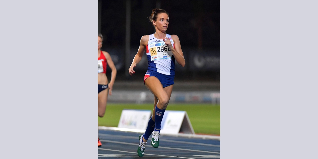 Lucy Evans running on an athletics tracks. Lucy is a white woman with dark hair. She wears blue shorts and white top. A competitor in red runs behind. The track is blue with a green field and crowd in the background.