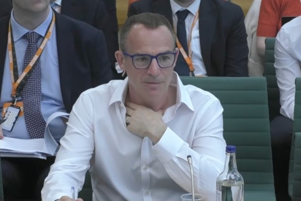 Martin Lewis sat in a green chair. Lewis is a white man with short dark hair, wearing glasses and a white shirt. He is surrounded by several people sat partially obscured either side of and behind him.