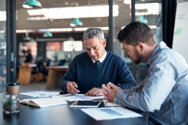 Two white men sit at a table analysing graphs. The man on the left is older with grey hair and a navy jumper. The man on the right has brown hair and beard and wears a light blue shirt.
