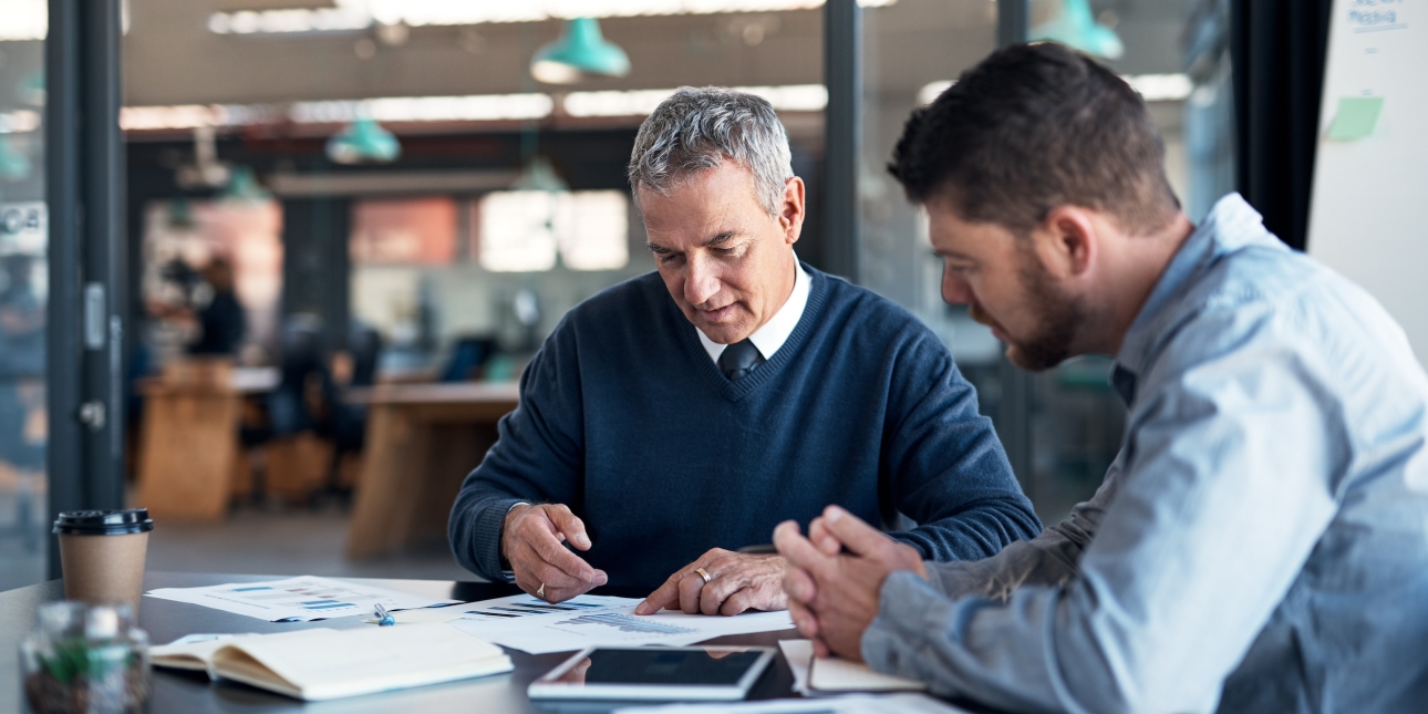 Two white men sit at a table analysing graphs. The man on the left is older with grey hair and a navy jumper. The man on the right has brown hair and beard and wears a light blue shirt.