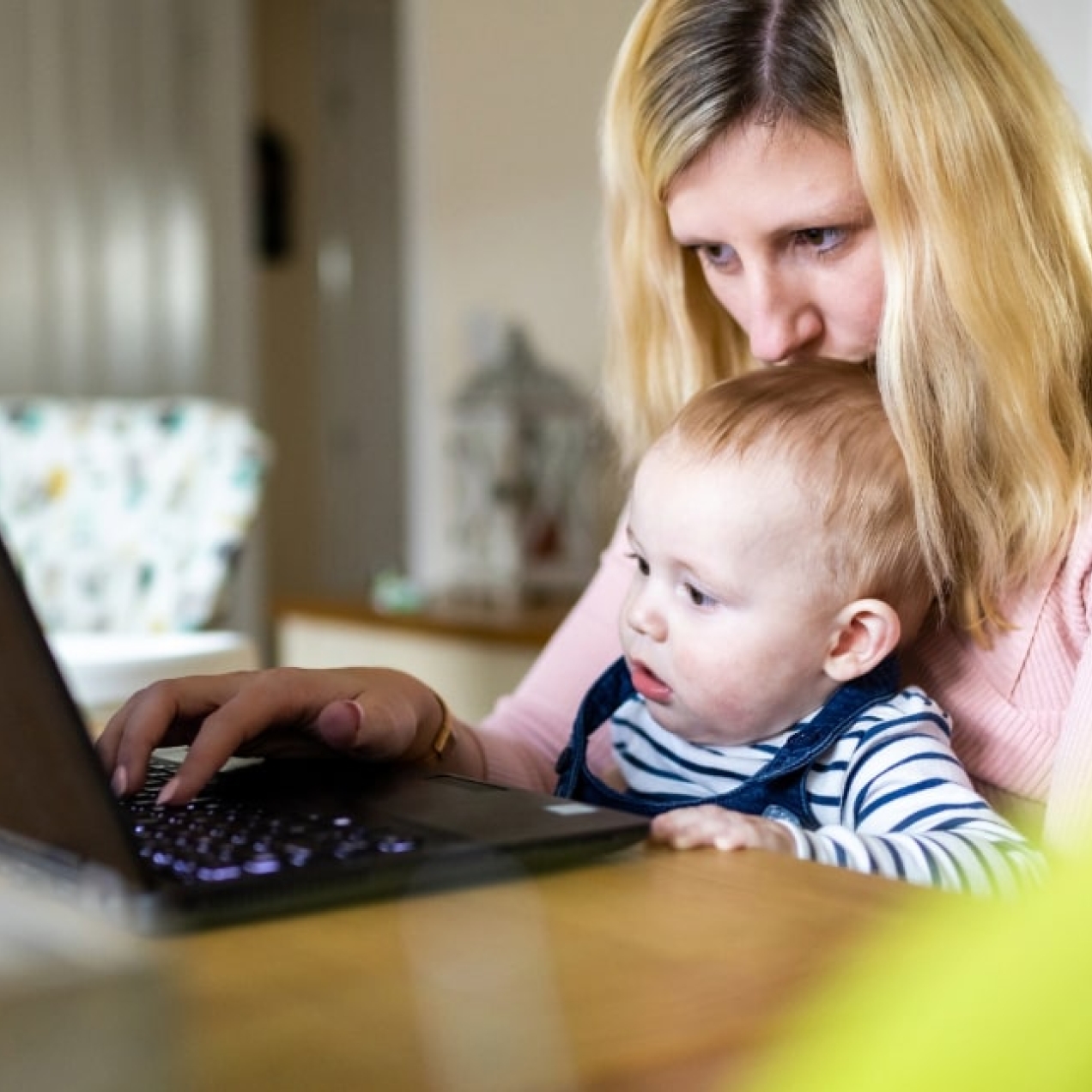At a kitchen table a white woman with blonde hair kisses her baby's head sat in her lap while working at her laptop
