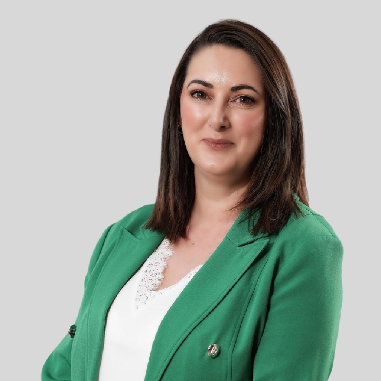 A colour portrait of Natasha Irvine against a grey background. Natasha is a white woman with shoulder length brown hair who wears a white top and green jacket. She smiles at the camera.