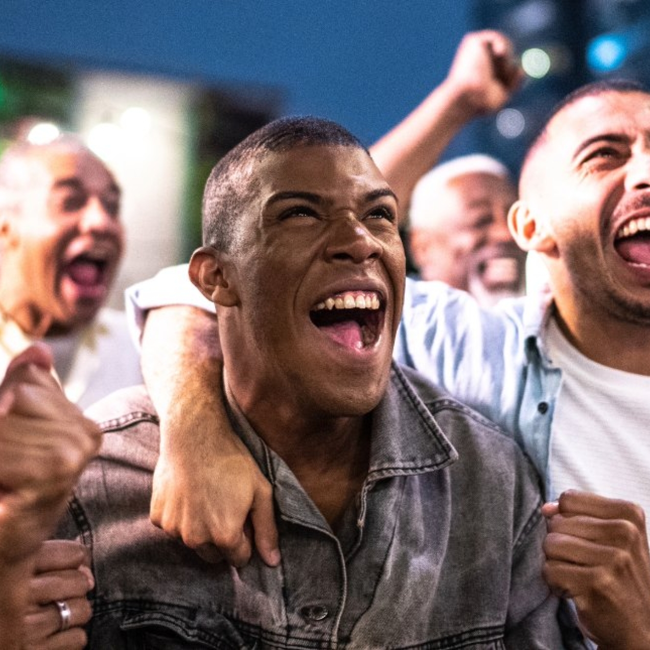 Three male friends cheering and celebrating likely sports action off-camera. Other people perform similar gestures in the background.