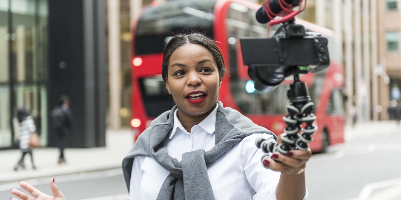 A woman in a London street filming herself talking at a camera with mounted microphone. It is attached to a handheld tripod which she holds in her left hand. The woman is black with a jumper tied over her shoulders over a white shirt. A red double de