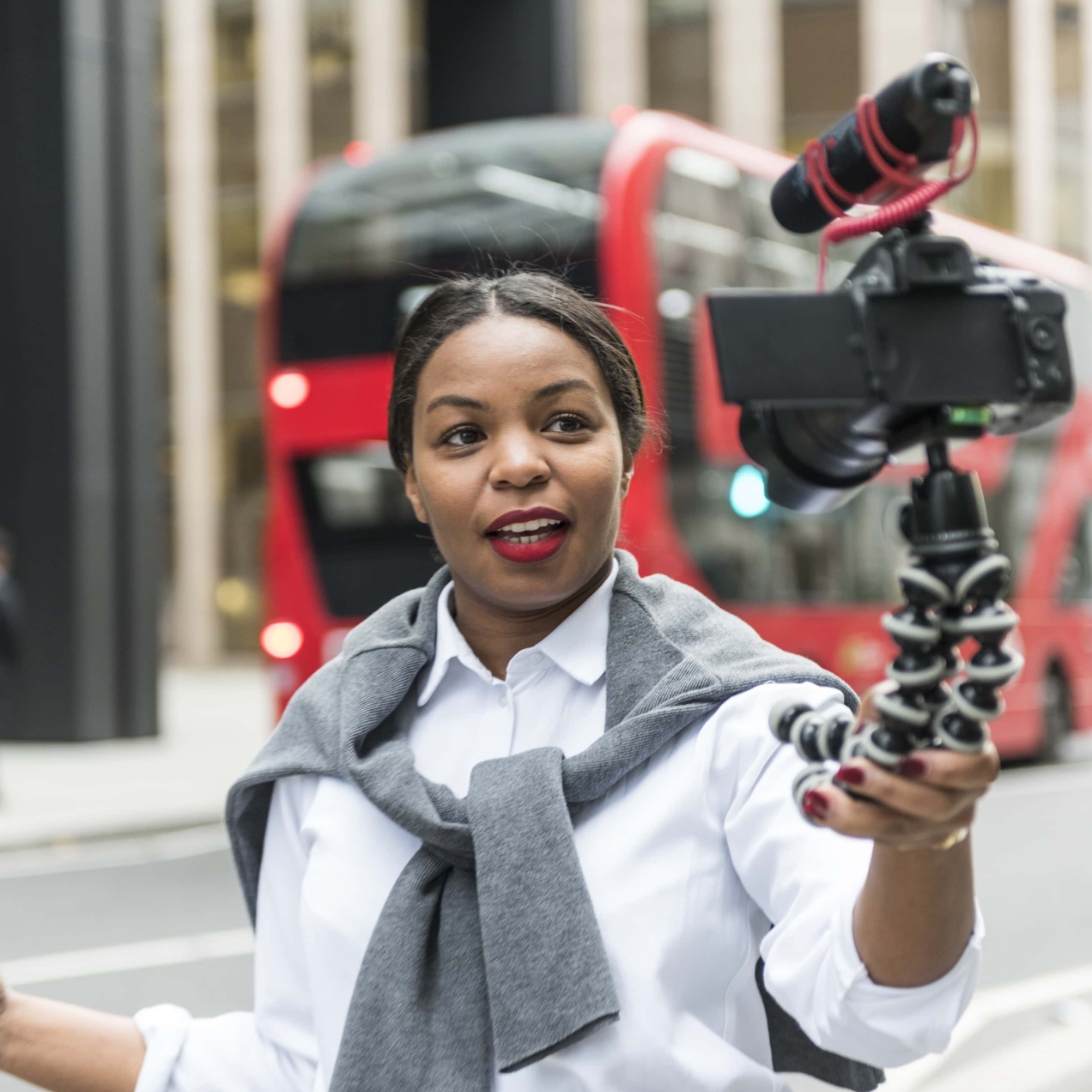 A woman in a London street filming herself talking at a camera with mounted microphone. It is attached to a handheld tripod which she holds in her left hand. The woman is black with a jumper tied over her shoulders over a white shirt. A red double de