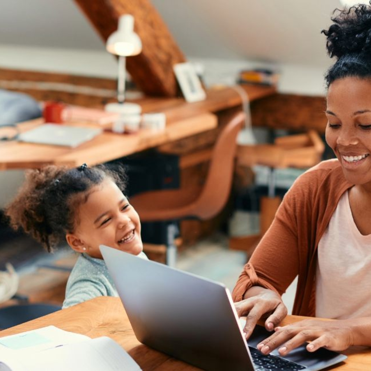 In a light filled home, a smiling Black woman wearing an orange cardigan works at her laptop as her child looks up at her smiling.