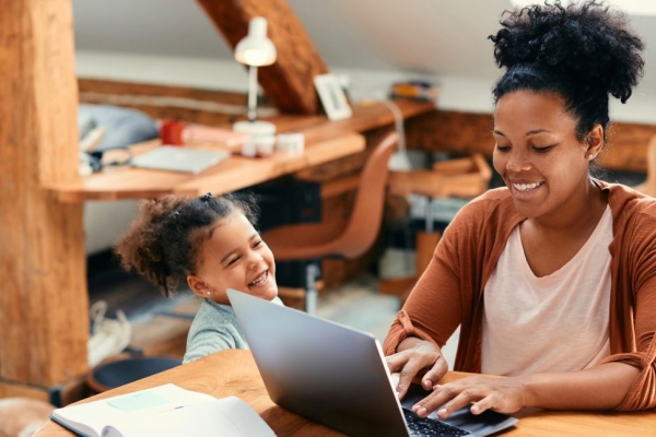 In a light filled home, a smiling Black woman wearing an orange cardigan works at her laptop as her child looks up at her smiling.