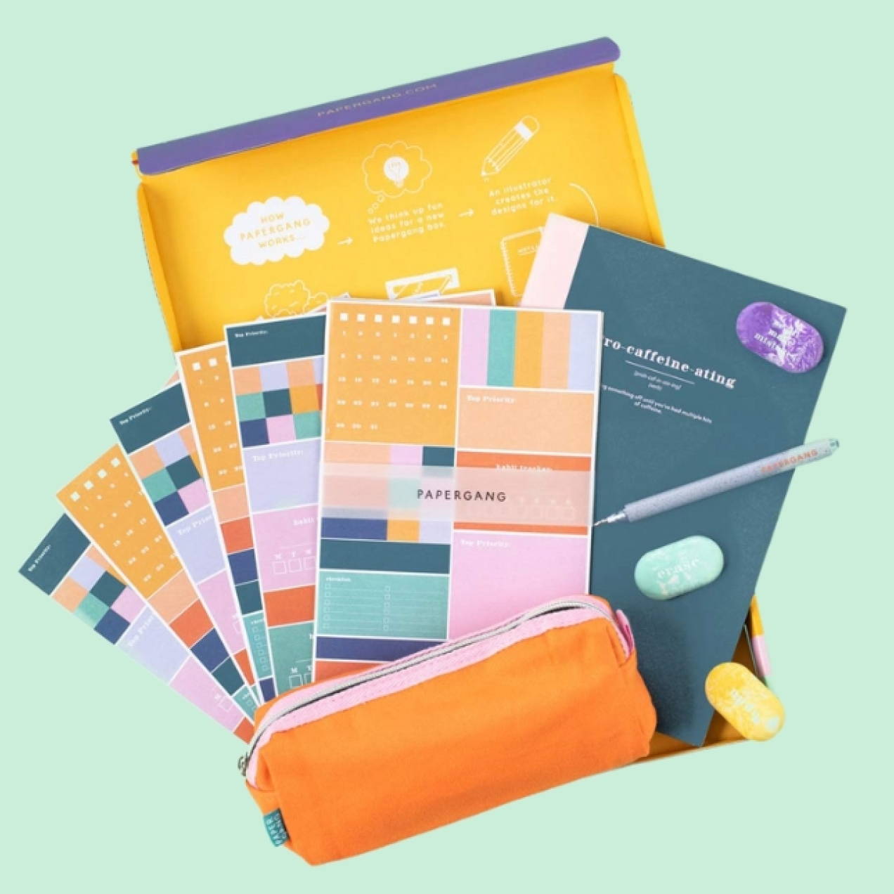 On a green pastel background, a birds eye view of an assortment of brightly coloured notebooks, planners, pencil case and other stationery
