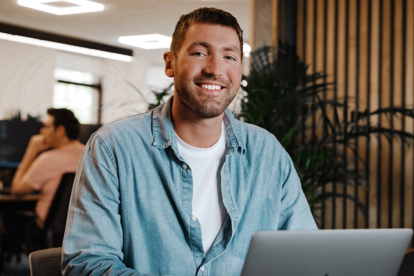 Ollie Manster, a white man with cropped brown hair, sat at a laptop smiling to camera. He wears a denim shirt over a white t-shirt, in modern office