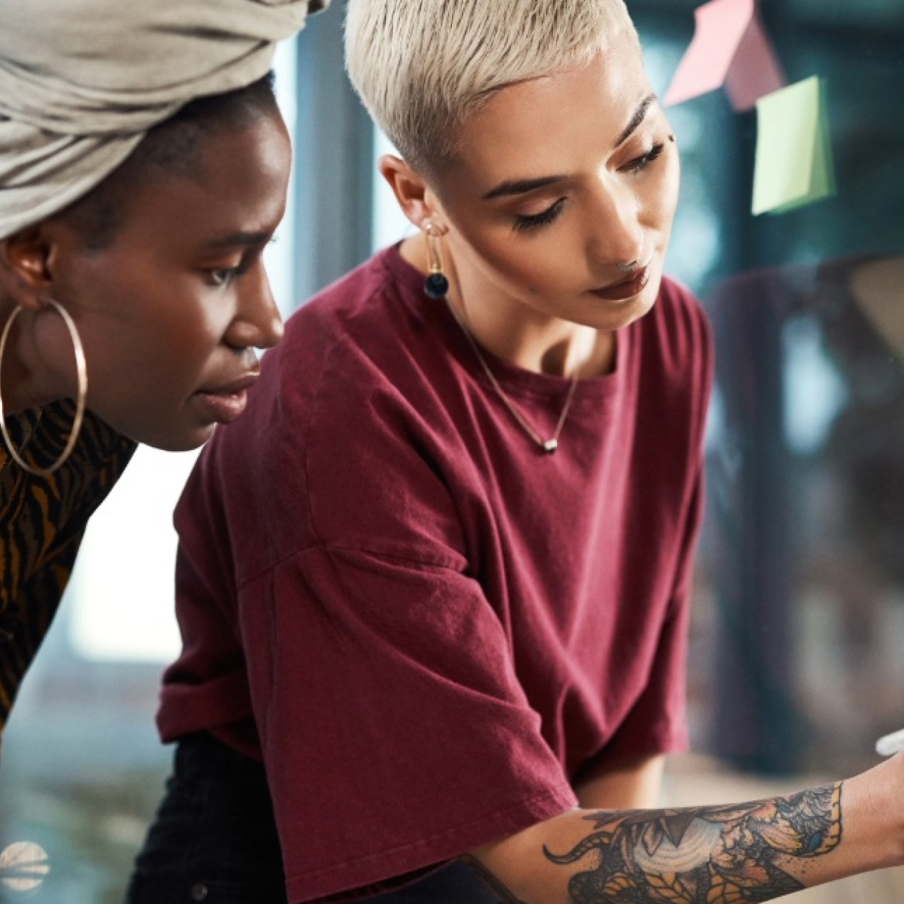 On the left of the photo, a Black woman wearing a grey head wrap looks on as her colleague - stood on the right a white woman with cropped, bleached hair and tattooed arm - writes on a sticky-note stuck to a glass wall. Both are bending down.