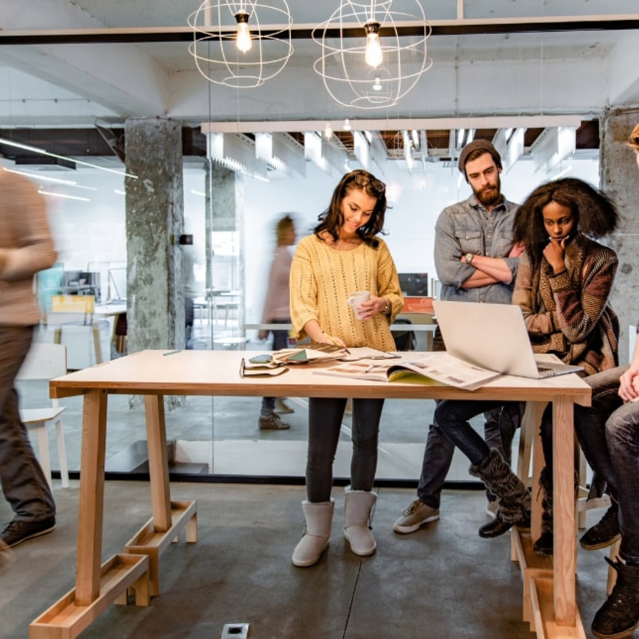 Two white men, a black woman and a white woman sit around a table looking at a laptop while other figures are blurred in the bacground walking past the glass office in which they are sat or stood