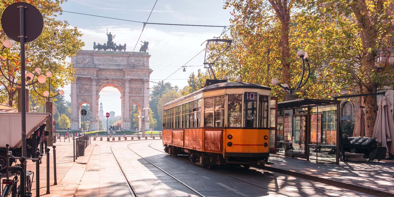 A vintage orange tram in the old town of Milan on a sunny day, with the Arco della Pace looming in the background.