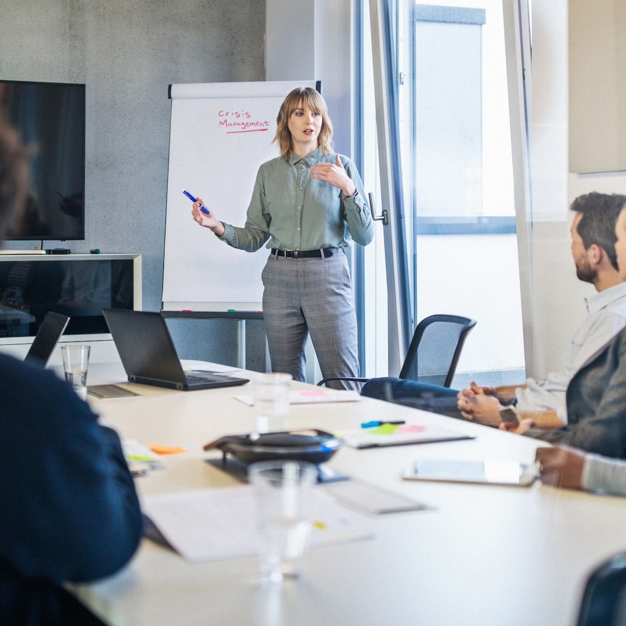 A white woman stands while addressing colleagues of different genders and ethnicities sat around a board table. Next to her is a display board with the words crisis management
