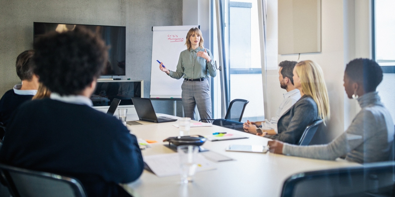 A white woman stands while addressing colleagues of different genders and ethnicities sat around a board table. Next to her is a display board with the words crisis management