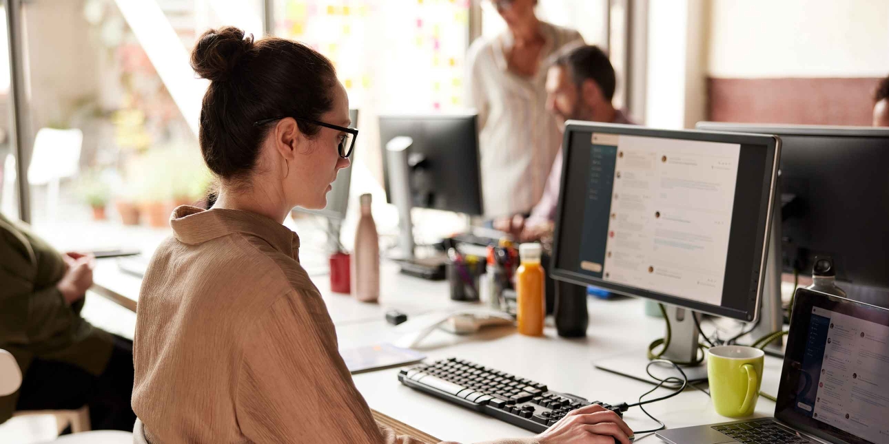 A side profile of a white woman sat at a desk holding a mouse in her right hand while looking at emails on her computer screen. She has dark hair in a bun, glasses and wears a light brown shirt. Three other unidentified people are in the background