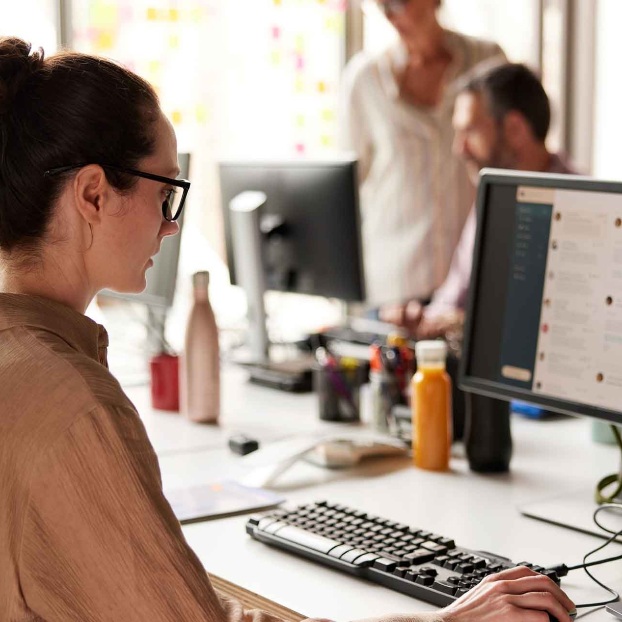 A side profile of a white woman sat at a desk holding a mouse in her right hand while looking at emails on her computer screen. She has dark hair in a bun, glasses and wears a light brown shirt. Three other unidentified people are in the background
