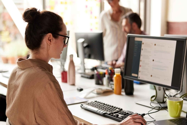 A side profile of a white woman sat at a desk holding a mouse in her right hand while looking at emails on her computer screen. She has dark hair in a bun, glasses and wears a light brown shirt. Three other unidentified people are in the background