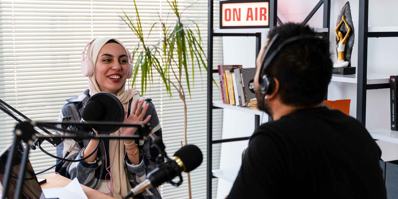 A smiling woman wearing large headphones over her cream hijab engages in conversation during a podcast. She faces an unidentified man with dark hair and black shirt. They are surrounded by microphones, laptop, notepad and an on-air sign upon a shelf.
