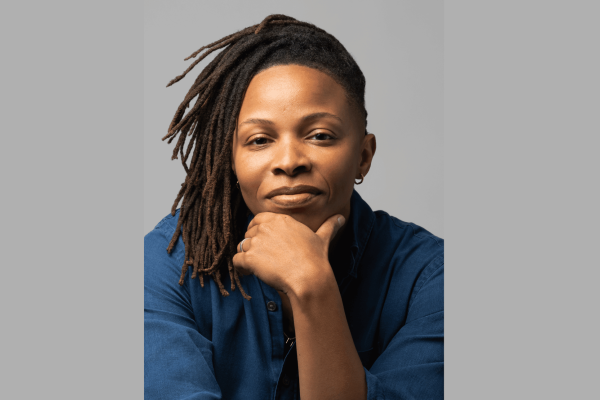 Prentis Hemphill, a black person with shoulder length hair and blue top, holds their chin in their left hand while looking at the camera