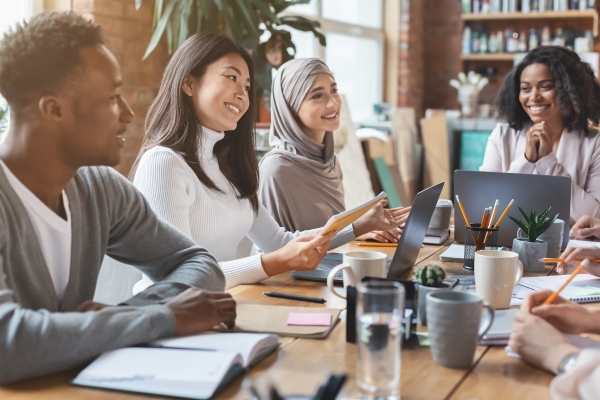 Smiling colleagues sat around a table with books, mugs and laptops. From left to right: a young Black man with short hair and grey jumper; a south-east Asian woman with shoulder length hair and white jumper; an Asian woman wearing a grey hijab; a Bla
