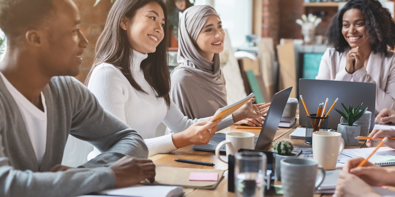 Smiling colleagues sat around a table with books, mugs and laptops. From left to right: a young Black man with short hair and grey jumper; a south-east Asian woman with shoulder length hair and white jumper; an Asian woman wearing a grey hijab; a Bla