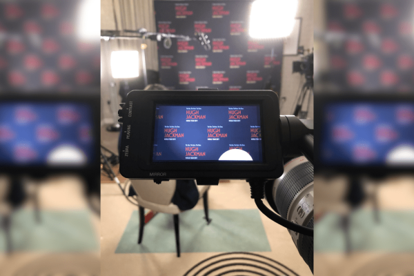 Looking through a video camera screen at the background of an empty chair, portable lights and boom microphone which are situated in front of a dark blue screen with red type which reads Hugh Jackman world tour