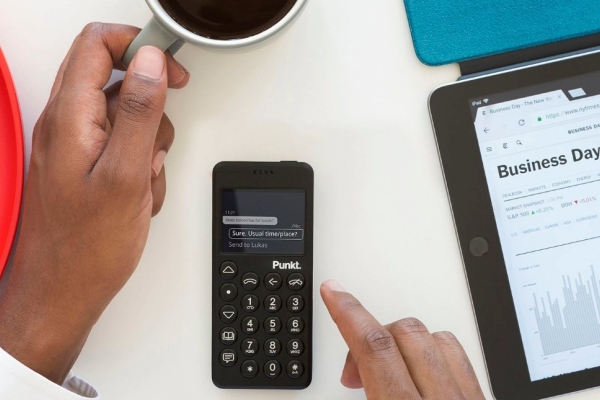 An unidentified right hand operates a Punkt device while a cup of coffee is held in the left hand. Adjacent is a tablet with a screen showing a business day calendar.