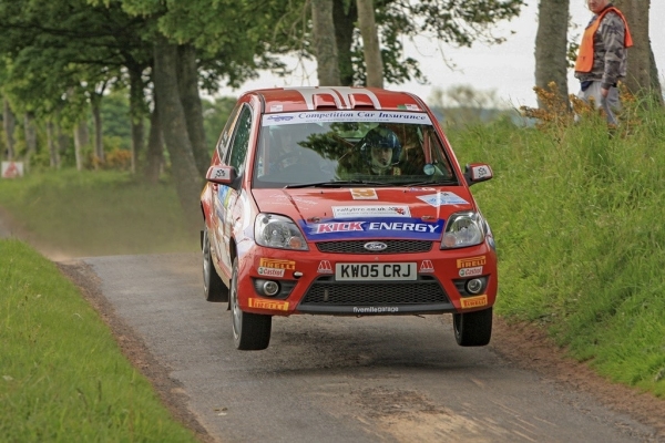 A red Ford Focus rally car emblazoned with sponsor logos takes to the air as it speeds down a narrow track with green verges either side