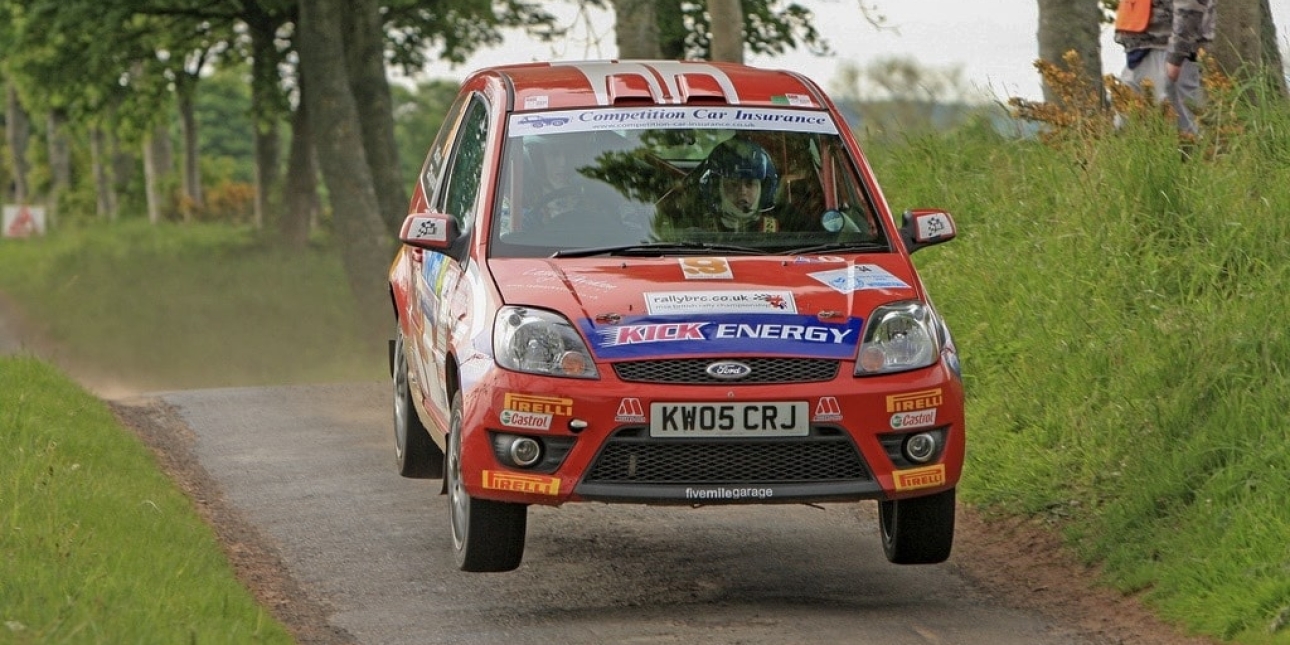 A red Ford Focus rally car emblazoned with sponsor logos takes to the air as it speeds down a narrow track with green verges either side
