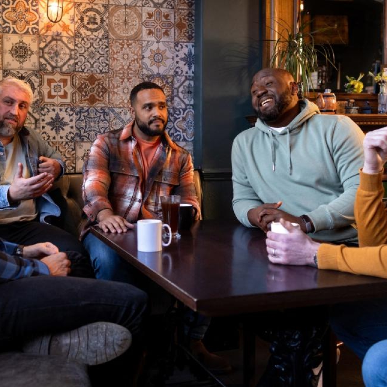 Five middle aged men sat chatting around a dark wooden table in a pub or cafe. The wall behind them features colourful, decorative tiles and a large mirror.