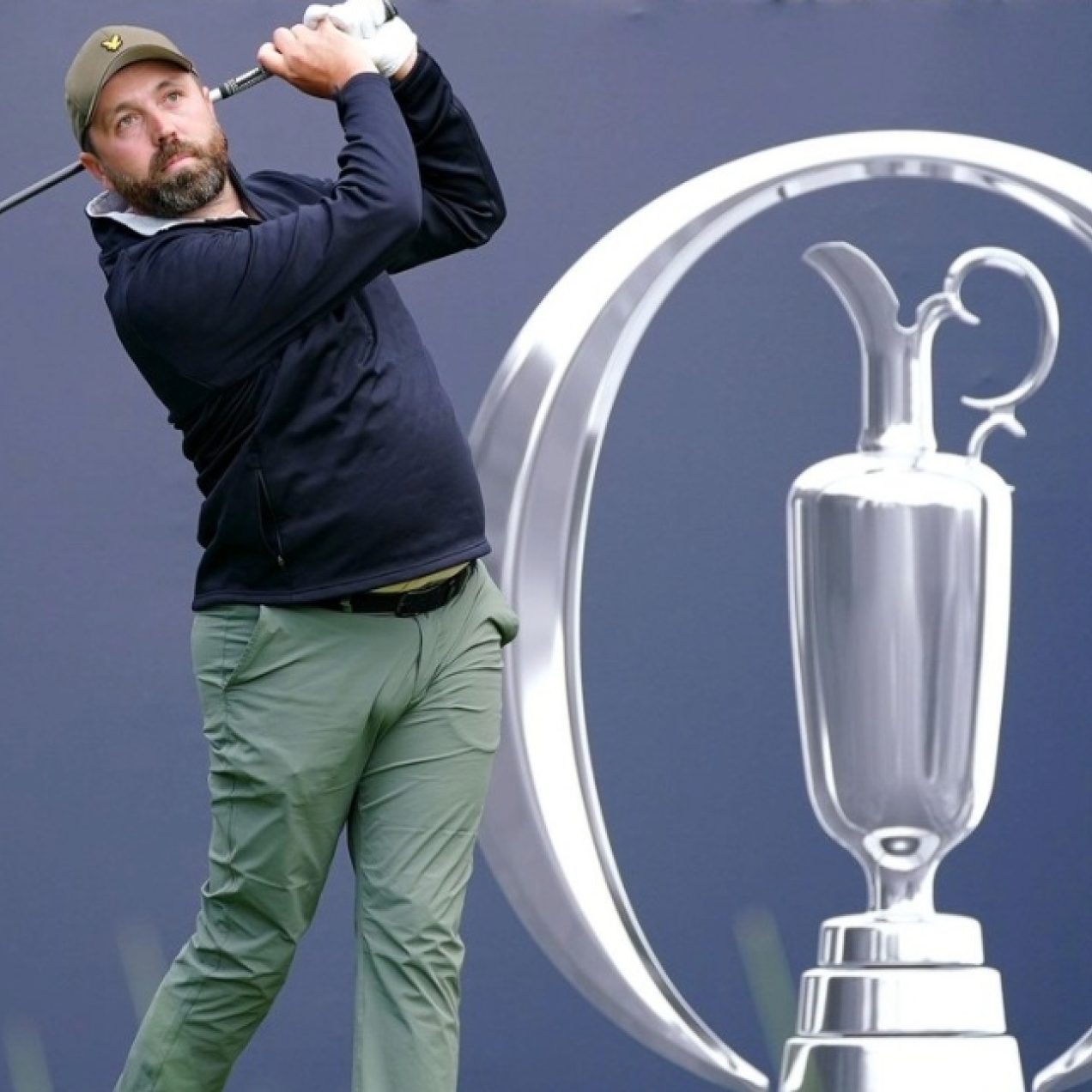 Golf influencer Rick Shiels tees off. He is a white man with a beard who wears a green cap, blue top and green trousers. He is in front of the a blue hoarding with a silver emblem.