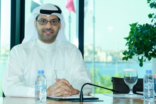 Saad Al Rubaiaan wearing glasses and a white ghutra and thobe while sat at a desk with water bottles and glasses on it. He is in front of a window which has flags and greenery outside