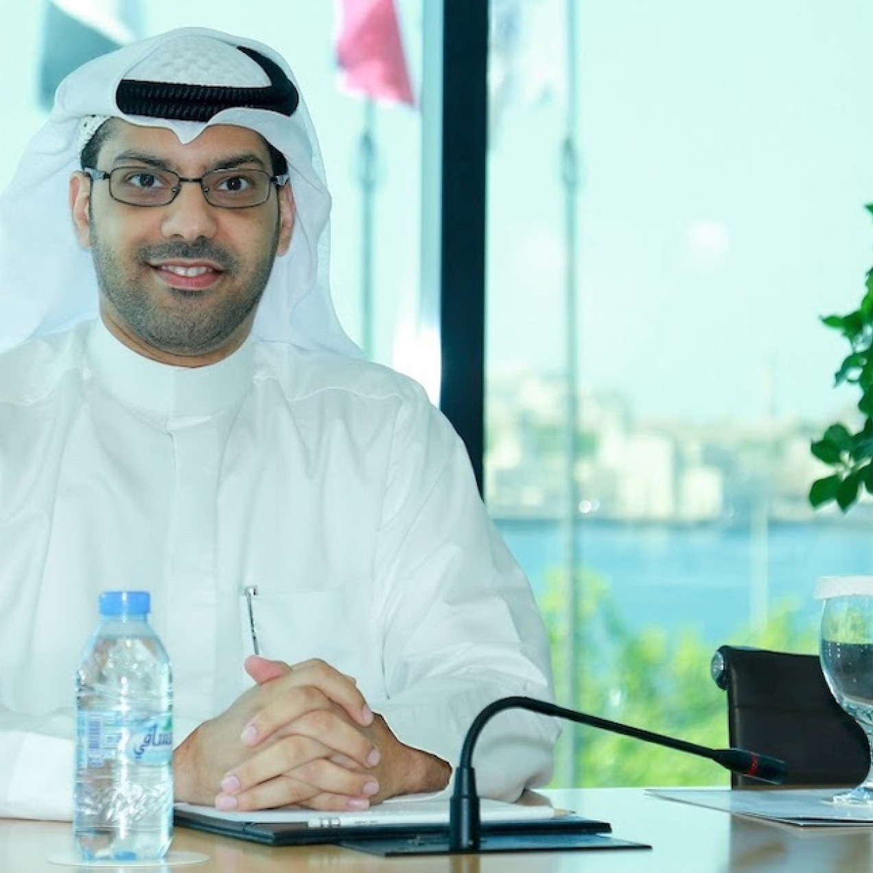 Saad Al Rubaiaan wearing glasses and a white ghutra and thobe while sat at a desk with water bottles and glasses on it. He is in front of a window which has flags and greenery outside