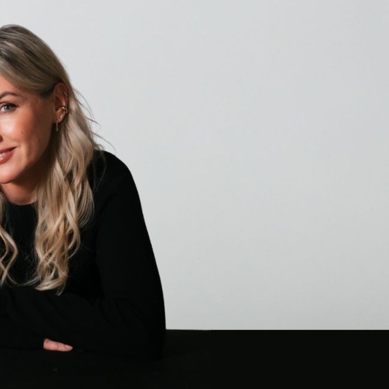 A colour portrait of Sali Hughes. Sali is a white woman with blond hair who is wearing black. She is sat leaning on a black desk against a grey background.