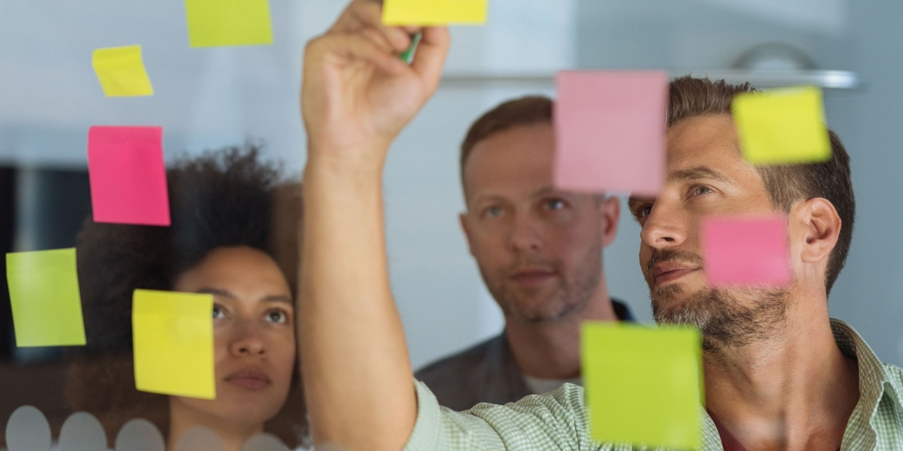Looking through a glass wall adorned with multicoloured sticky notes at three colleagues - a Black woman and two white men. The man on the right is writing on one of the notes as the other look on.