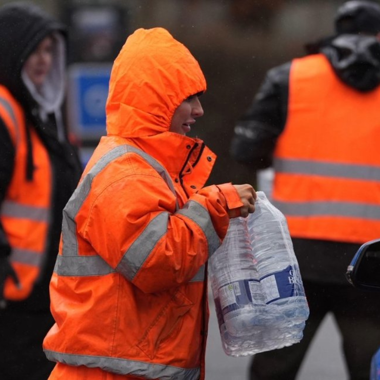A worker in orange hi-vis clothing hands over bottled water to unseen recipients of a blue car.