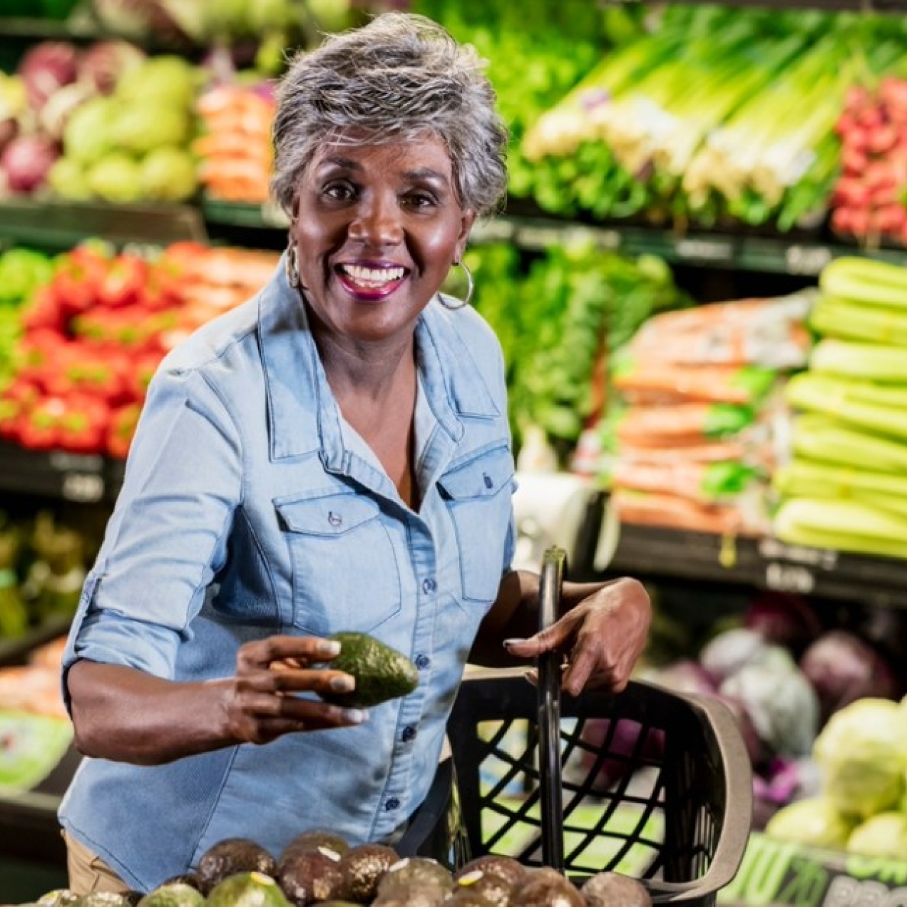 In a supermarket aisle surrounded by displays of fruit and vegetables a mature, smiling Black woman with short grey hair reaches for avocados.