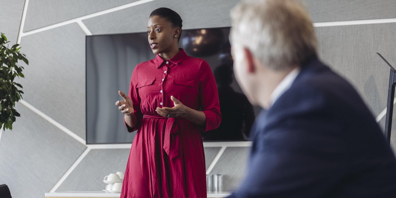 A Black woman with short hair, red dress and her arms open addresses a meeting. Sat to her left is an unidentified white male in a blue suit. The walls are grey patterned and there is a screen behind her.