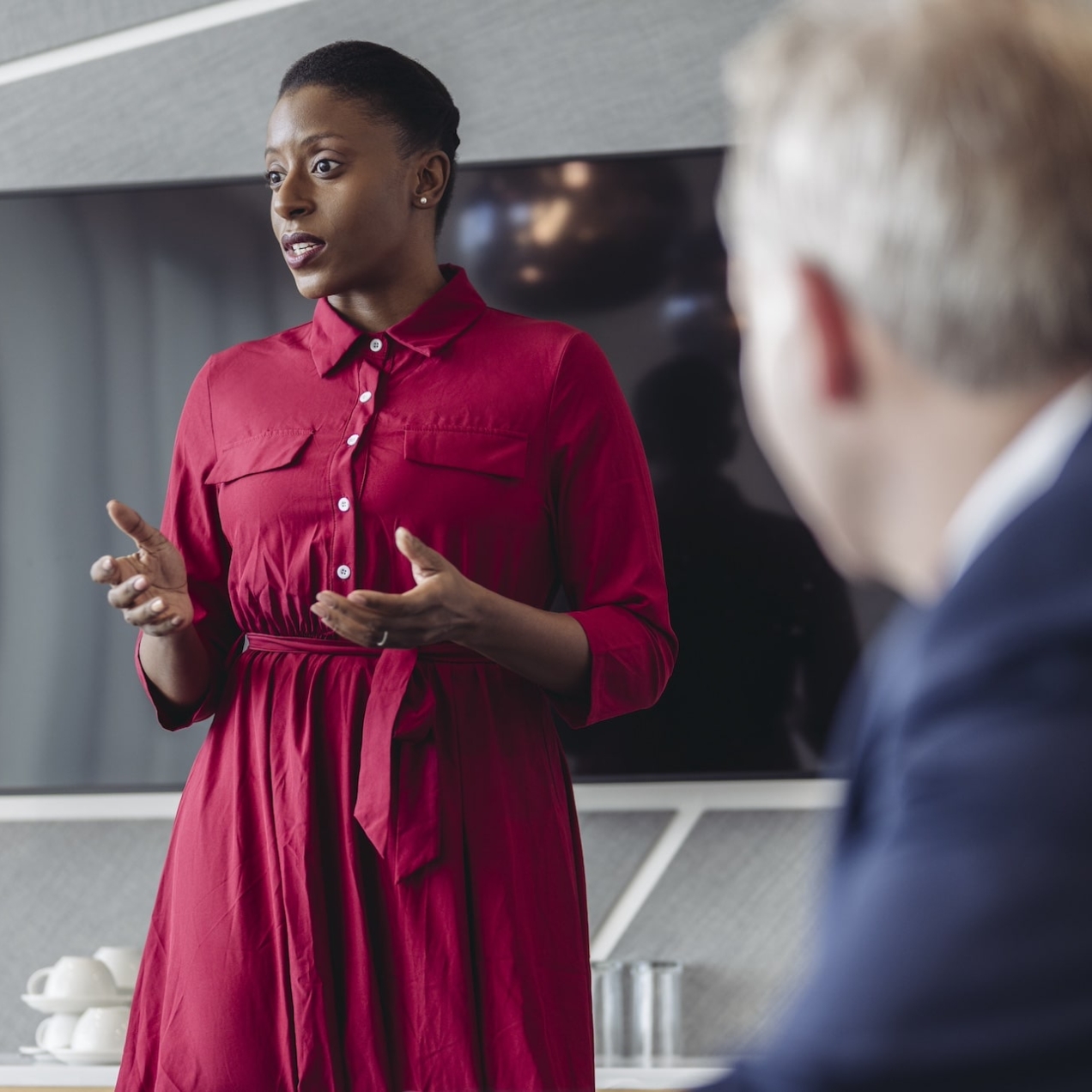 A Black woman with short hair, red dress and her arms open addresses a meeting. Sat to her left is an unidentified white male in a blue suit. The walls are grey patterned and there is a screen behind her.