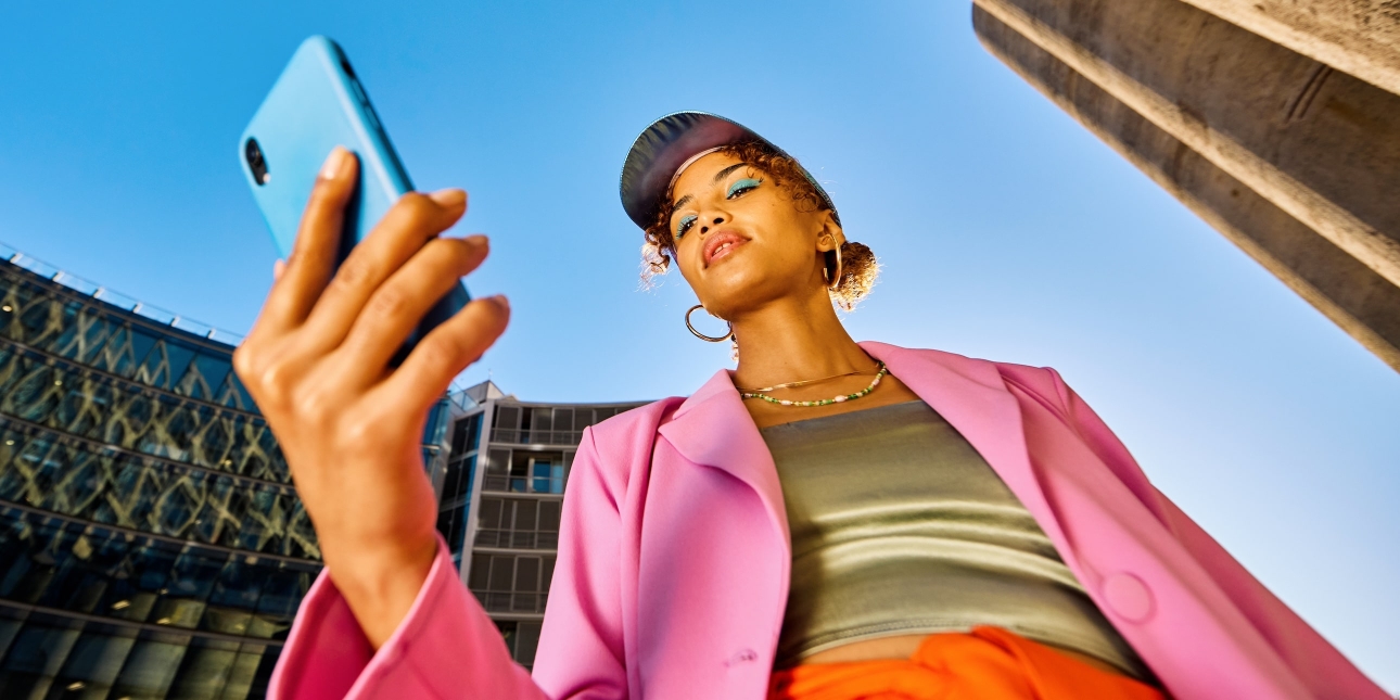 A low angle shot of a Black woman holding a turquoise phone against a bright blue sky. She wears blue eye shadow, a cap, bright pink jacket, green shimmering top and orange skirt. There are buildings on either side