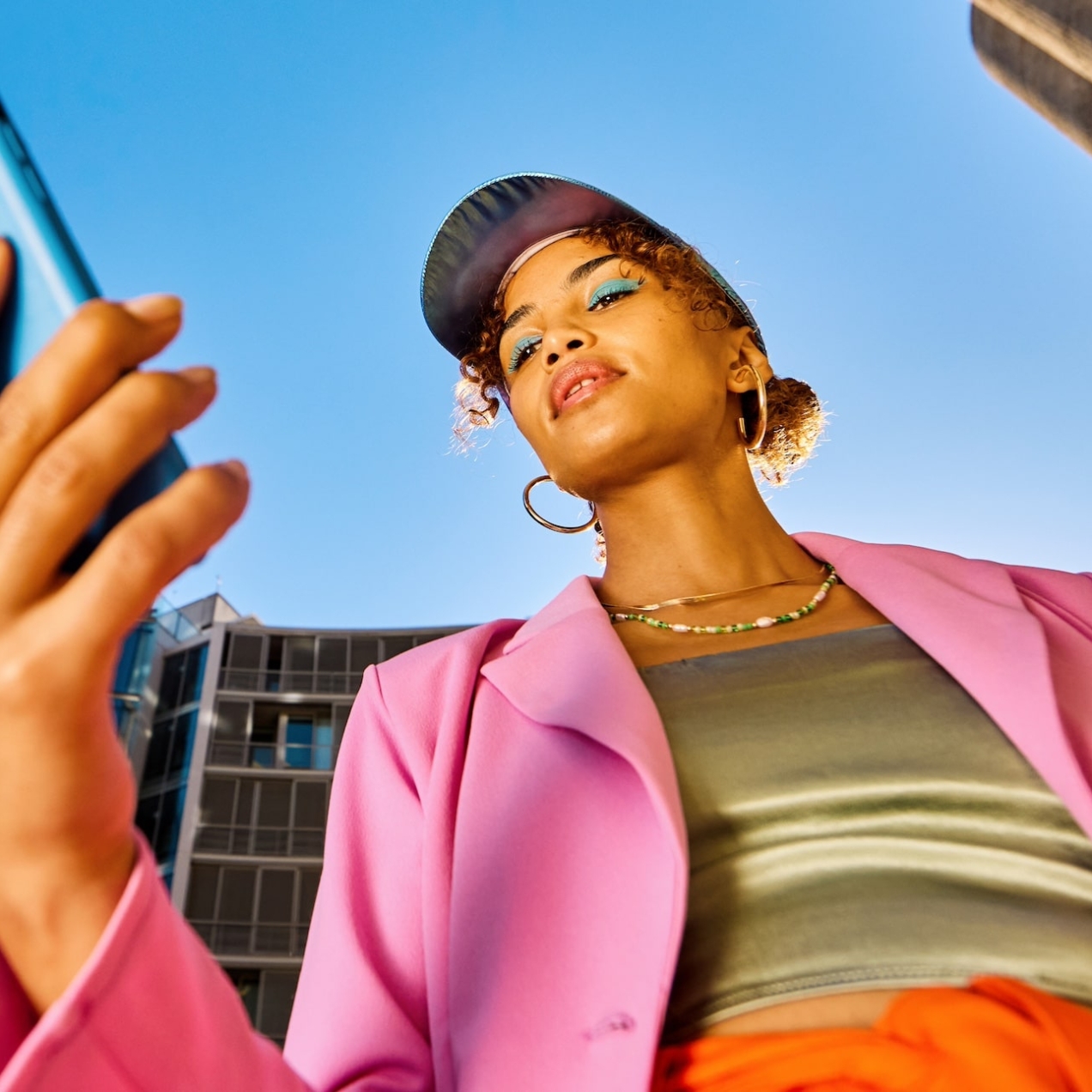 A low angle shot of a Black woman holding a turquoise phone against a bright blue sky. She wears blue eye shadow, a cap, bright pink jacket, green shimmering top and orange skirt. There are buildings on either side