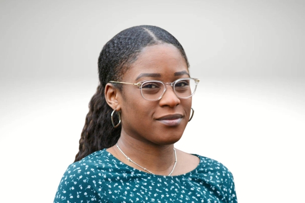 A portrait of Tani Fatuga, a Black woman with long dark hair and glasses, who wears a blue patterned top while looking at the camera. The background is grey-white gradient.