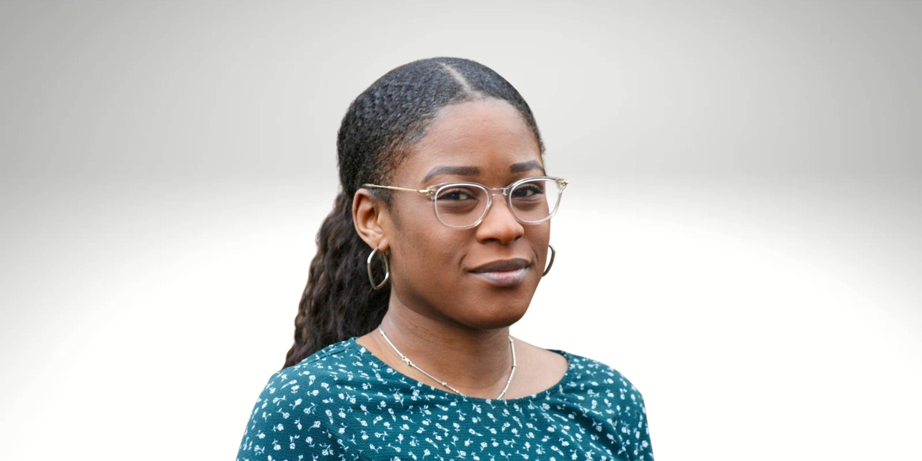 A portrait of Tani Fatuga, a Black woman with long dark hair and glasses, who wears a blue patterned top while looking at the camera. The background is grey-white gradient.