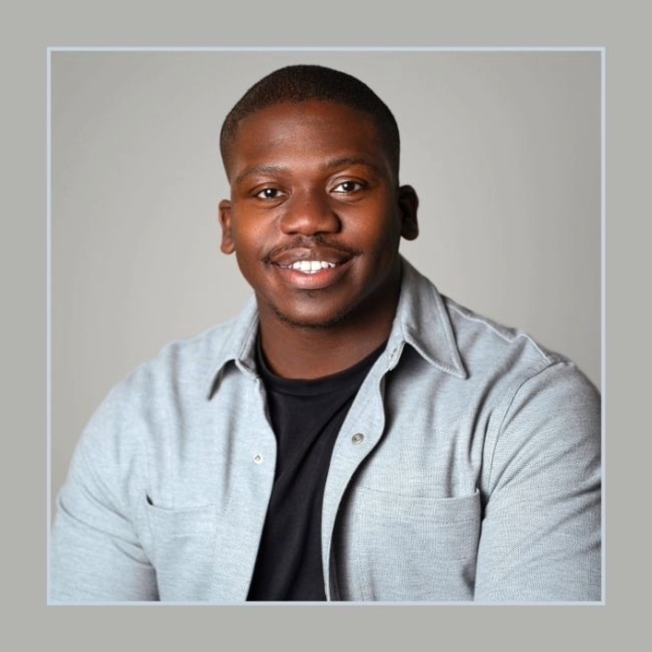 A portrait of Tariq Peters on a grey background. Tariq is a Black man with short hair and a light-grey shirt over a black t-shirt