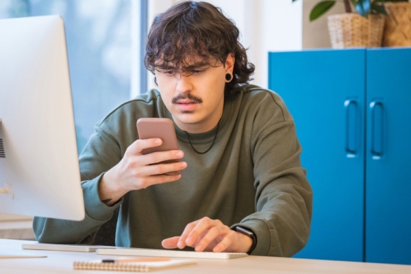 A young white man with dark hair and moustache grimaces as he stares at the mobile in his hand. He is sat in a front of a computer at a desk with a blue cabinet to the right of the image.