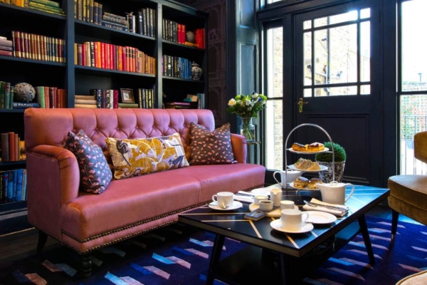 A dark blue room with a floor to ceiling bookcase, pink sofa and tan armchairs set around a coffee table with a traditional cream tea.