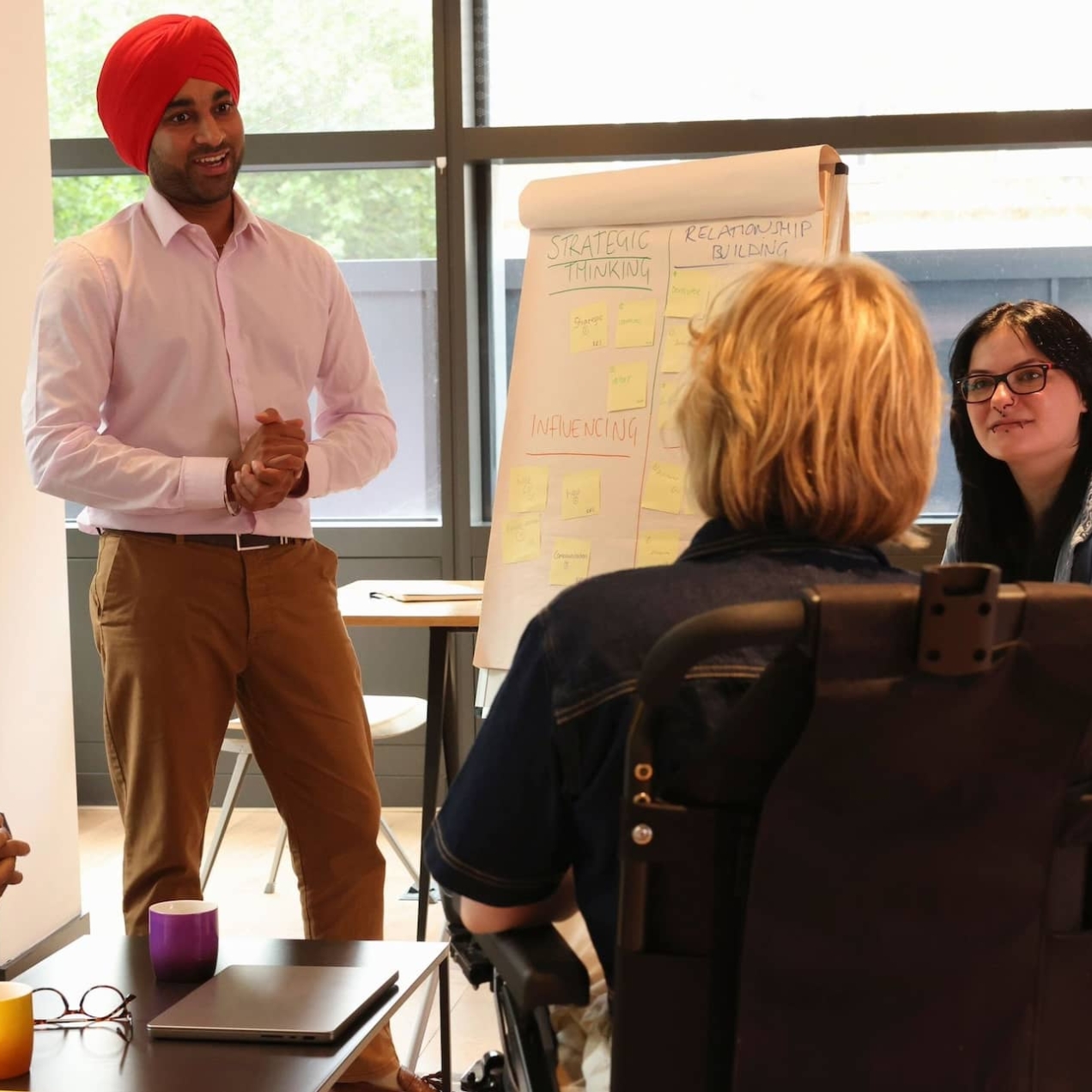 A group of five colleagues: A Black man in a blue shirt sat on a sofa. To his left stands an Asian man with an orange turban and pink shirt stood next to a flip chart. He is smiling at a person with blonde hair who uses a motorised wheelchair. To the