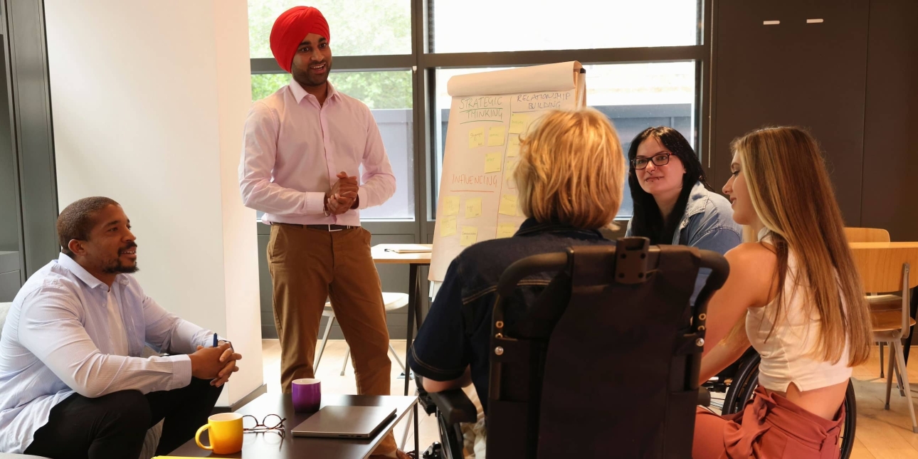 A group of five colleagues: A Black man in a blue shirt sat on a sofa. To his left stands an Asian man with an orange turban and pink shirt stood next to a flip chart. He is smiling at a person with blonde hair who uses a motorised wheelchair. To the
