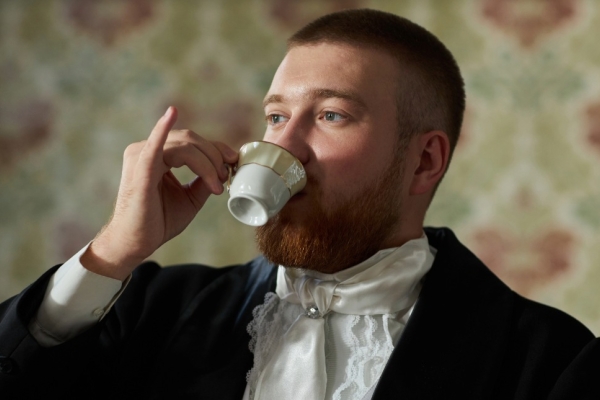 A white man with cropped brown hair and wearing a formal dark jacket and white ruffled shirt holds a porcelain tea cup with his pinkie finger extended.
